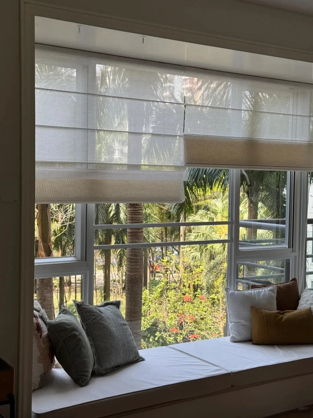 Cozy window seat with white cushions and assorted pillows, sheer blinds, palm trees visible outside