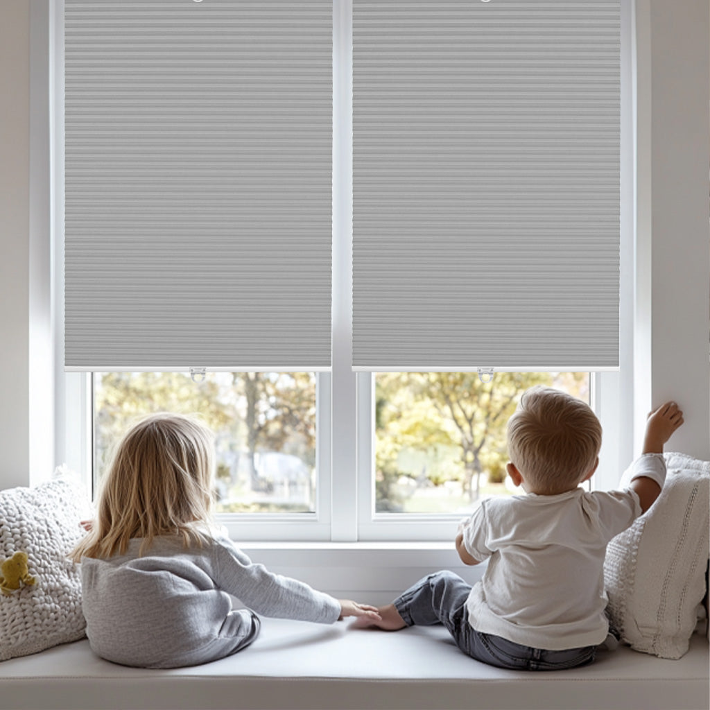 Two children sitting on a window seat with gray cellular shades partially closed and autumn view outside