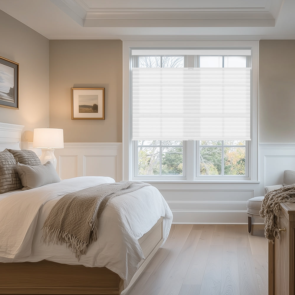 Cozy bedroom with white bedding, beige throw, wood furniture, table lamp, and large window with white blinds