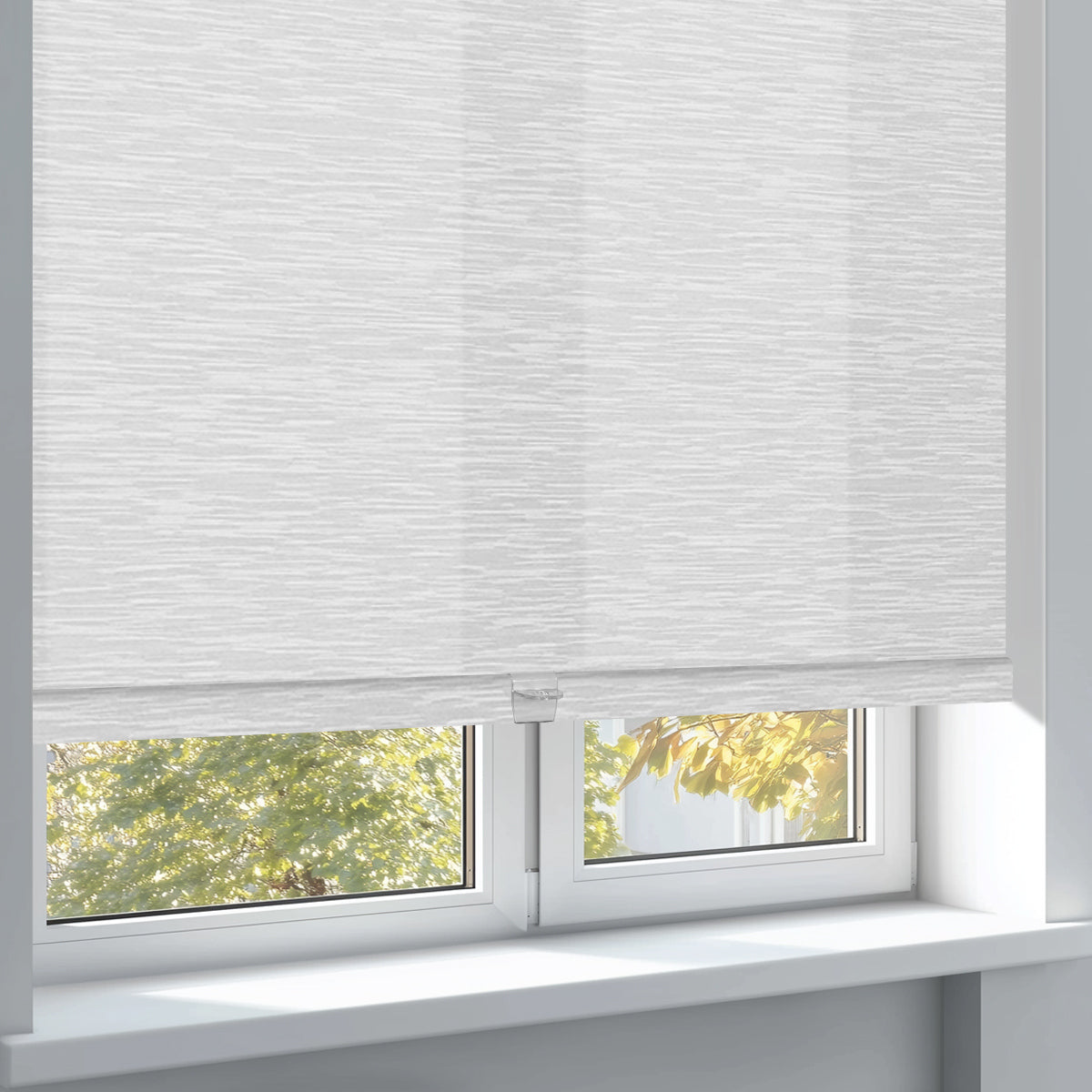 White textured roller blinds partially covering a window with green and yellow foliage outside